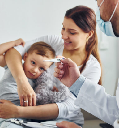 A doctor in a white coat and face mask holds a thermometer, showing it to a concerned mother holding her young child, who is resting his head on her shoulder in a clinic setting.