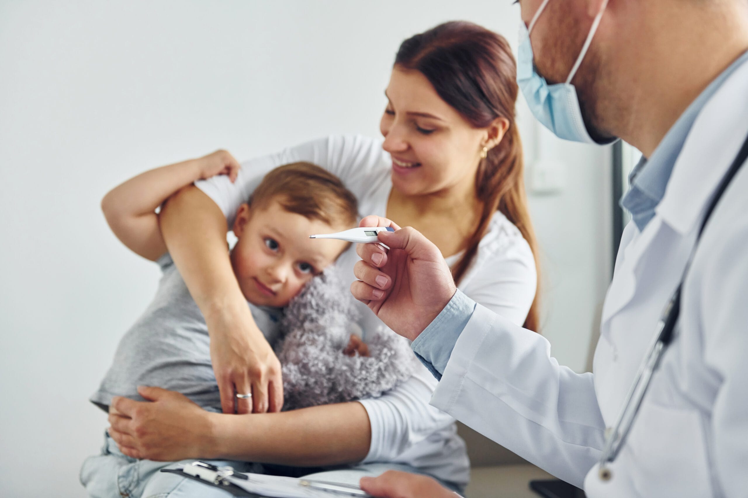 A doctor in a white coat and face mask holds a thermometer, showing it to a concerned mother holding her young child, who is resting his head on her shoulder in a clinic setting.