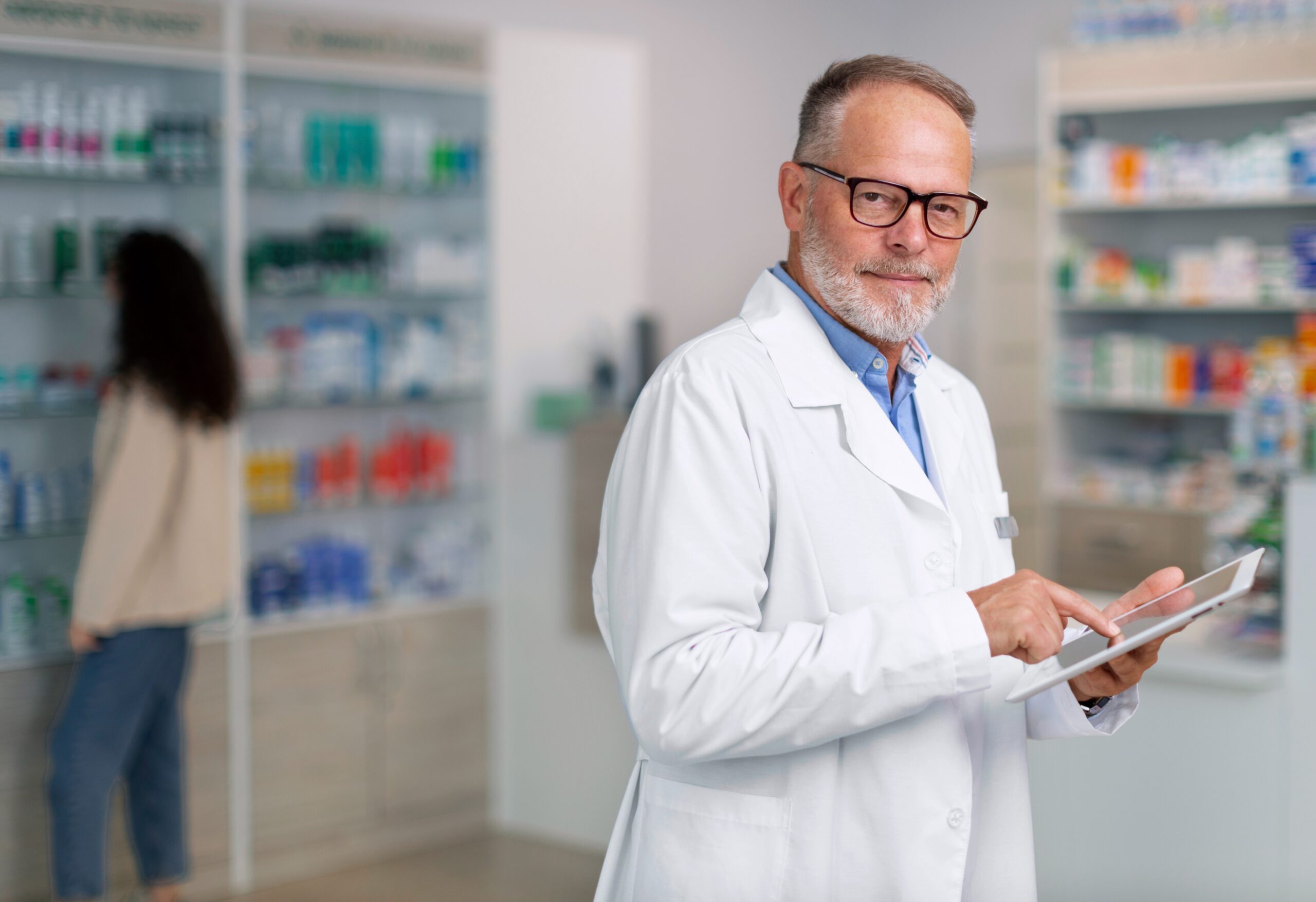 Pharmacist in a pharmacy, holding a tablet and reviewing medication records.