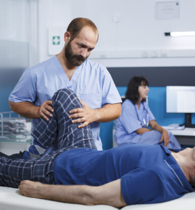 A healthcare professional examining an elderly patient's knee, with other medical staff in the background.