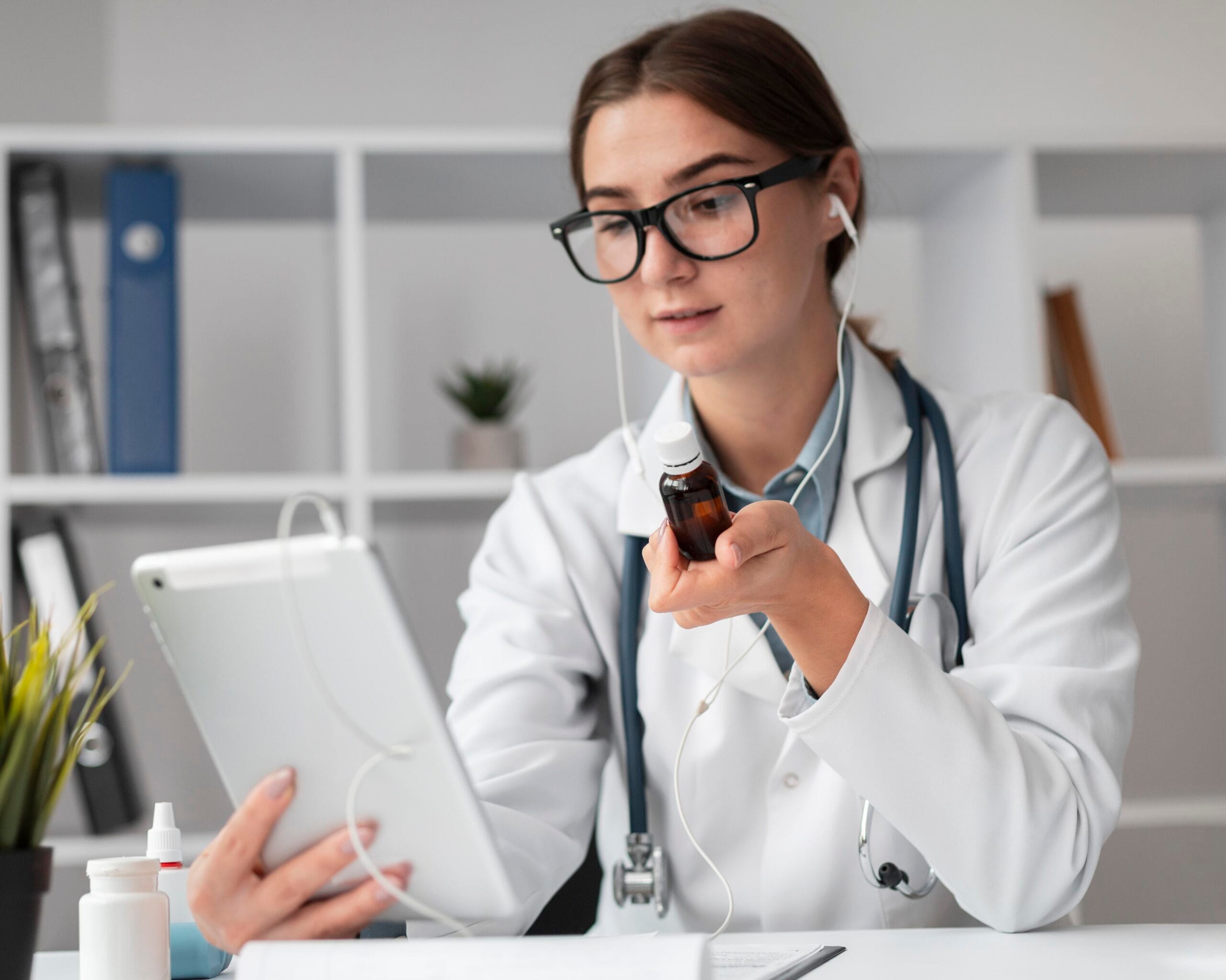 A telemedicine provider wearing headphones and a stethoscope, holding a medication bottle while reviewing notes on a tablet during a virtual consultation.