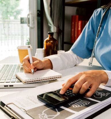 A healthcare professional in scrubs working at a desk, writing in a notebook while using a calculator, with a laptop and medical documents in the background.