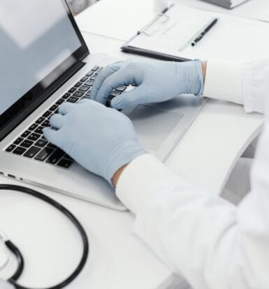 Healthcare professional wearing gloves typing on a laptop with a stethoscope nearby, representing medical scribing for accurate documentation.