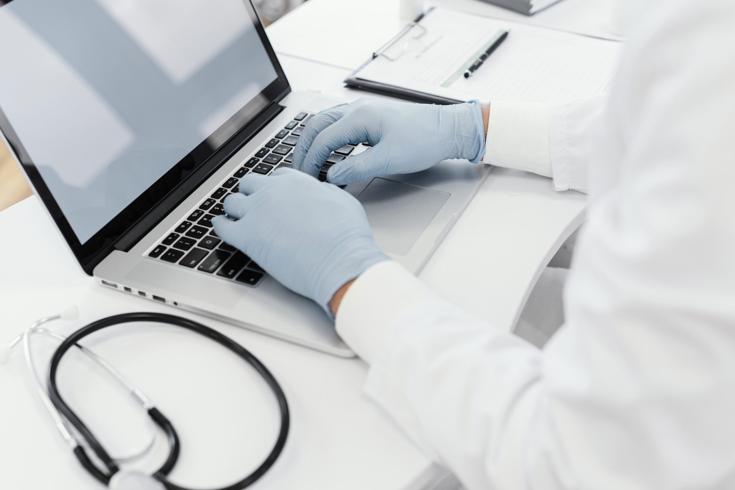 Healthcare professional wearing gloves typing on a laptop with a stethoscope nearby, representing medical scribing for accurate documentation.