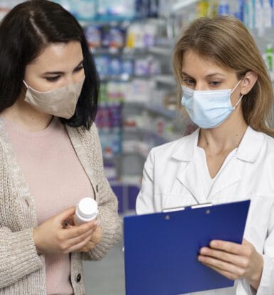 A pharmacist and a patient wearing masks discussing medication in a pharmacy. The pharmacist is holding a clipboard, and the patient is holding a prescription bottle.