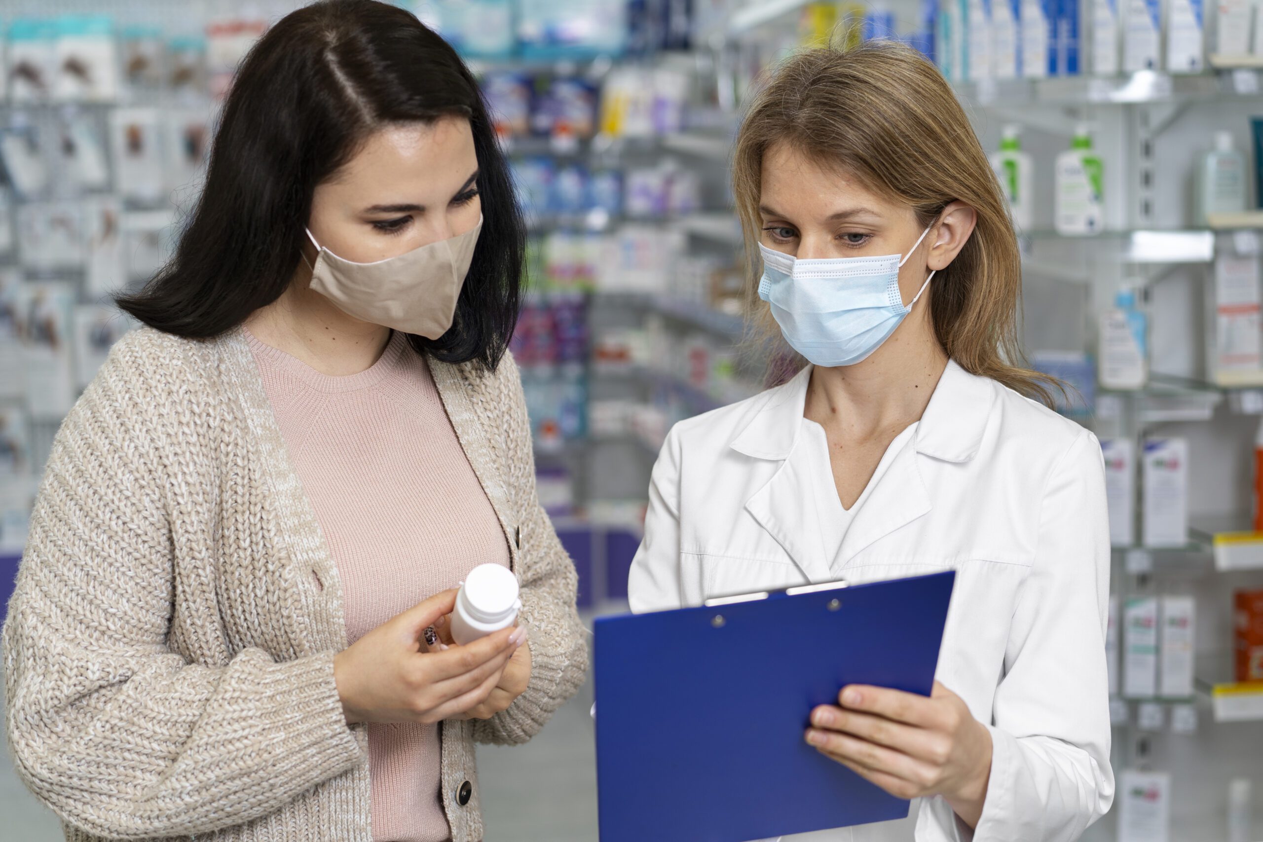 A pharmacist and a patient wearing masks discussing medication in a pharmacy. The pharmacist is holding a clipboard, and the patient is holding a prescription bottle.