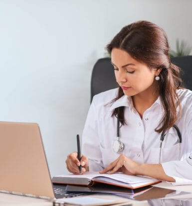 Female doctor or medical professional reviewing patient billing and coding information on a laptop while taking notes in a notebook