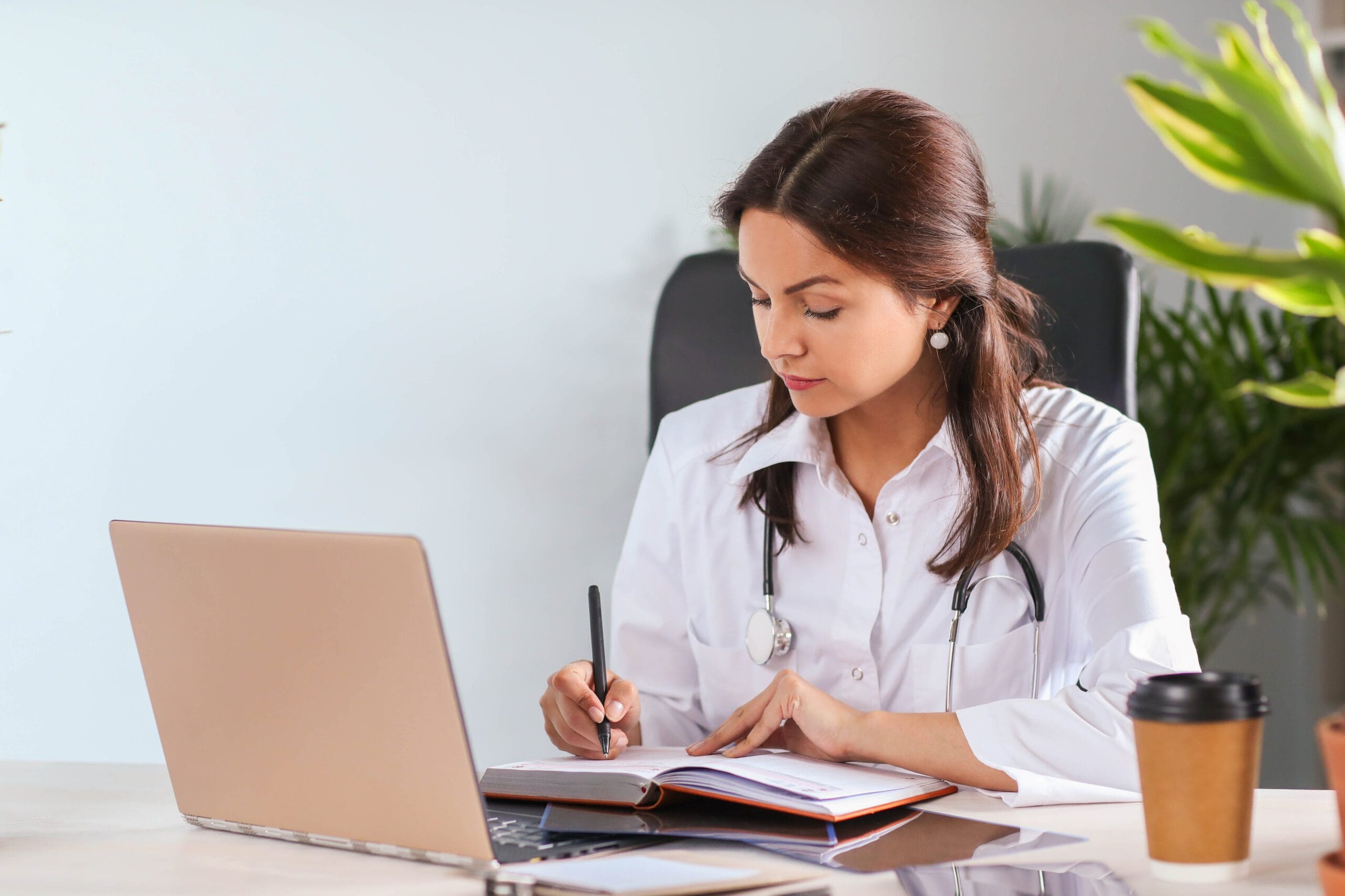 Female doctor or medical professional reviewing patient billing and coding information on a laptop while taking notes in a notebook