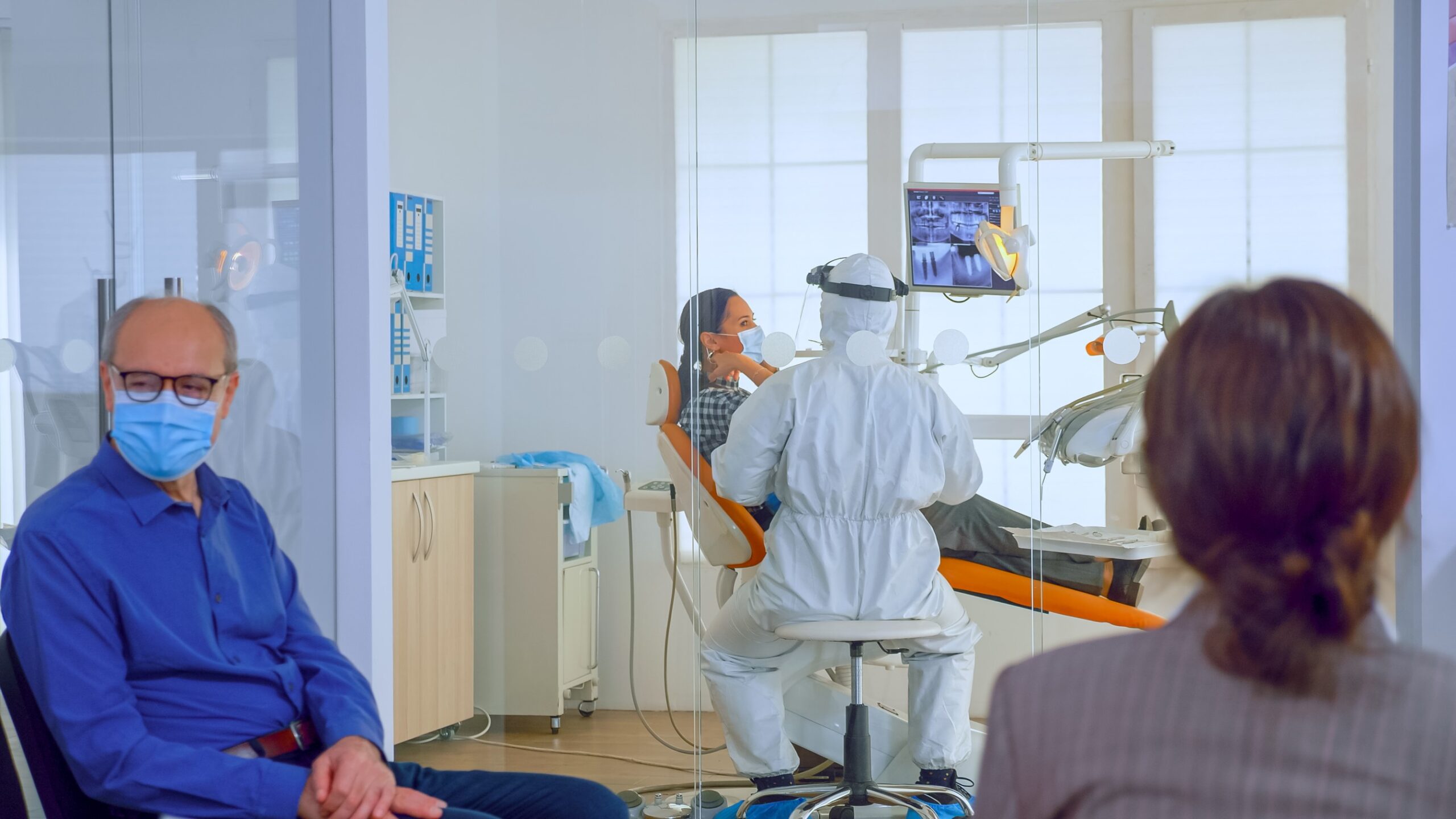 Healthcare provider in protective gear assisting a patient in a dental office, with another masked individual waiting in the background.