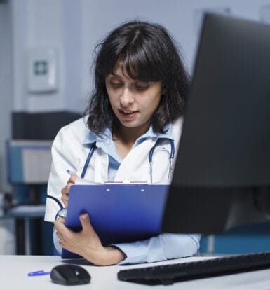 A healthcare professional with a stethoscope around her neck is reviewing a clipboard while sitting at a desk in front of a computer, possibly working on billing or administrative tasks in a long-term care setting.