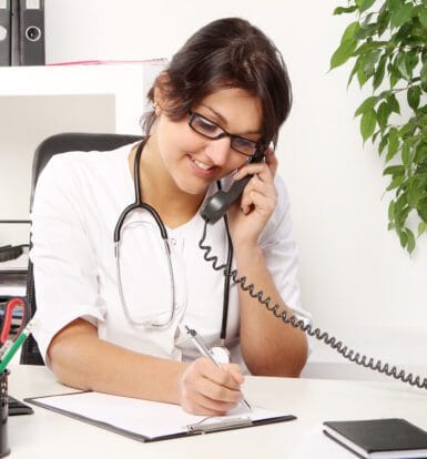 "Female healthcare professional wearing a stethoscope, talking on the phone while writing notes at her desk, representing a pharmacy call center providing patient support."