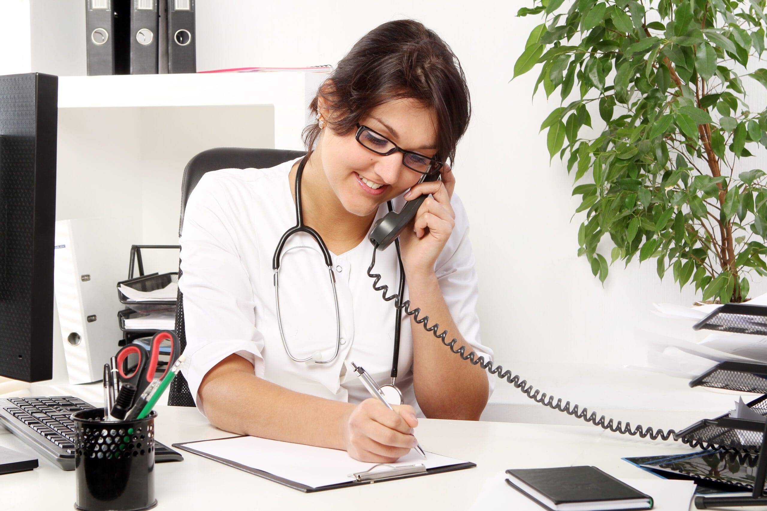 "Female healthcare professional wearing a stethoscope, talking on the phone while writing notes at her desk, representing a pharmacy call center providing patient support."