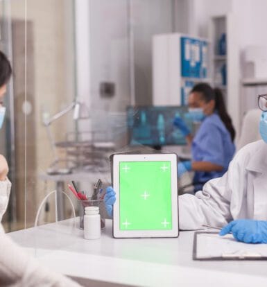 A doctor wearing a mask and gloves showing a patient a tablet screen in a clinical setting, while discussing medication or treatment options.