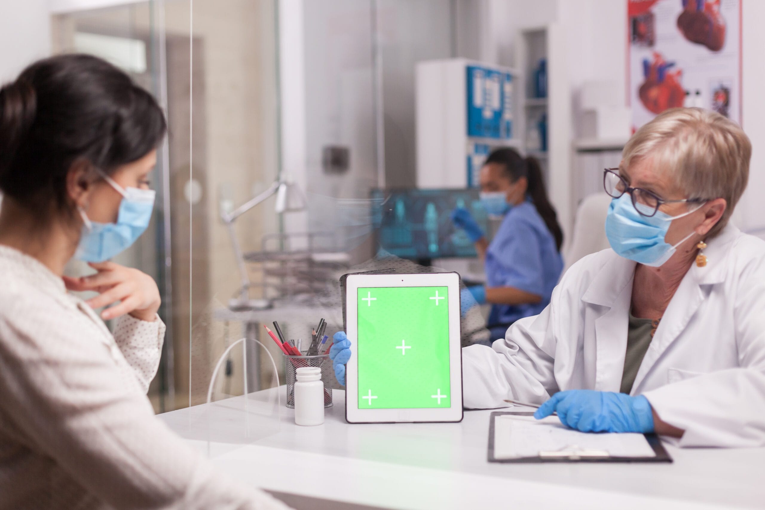 A doctor wearing a mask and gloves showing a patient a tablet screen in a clinical setting, while discussing medication or treatment options.