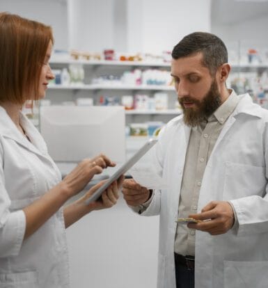 Two pharmacists discussing medication orders in a pharmacy, reviewing a tablet and a prescription for accuracy and drug utilization.