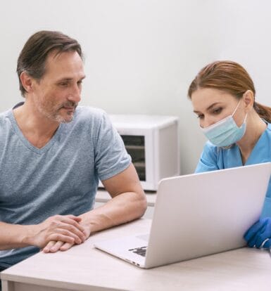 A healthcare provider in a blue uniform and face mask reviewing information on a laptop with a patient sitting beside her.