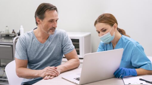 A healthcare provider in a blue uniform and face mask reviewing information on a laptop with a patient sitting beside her.