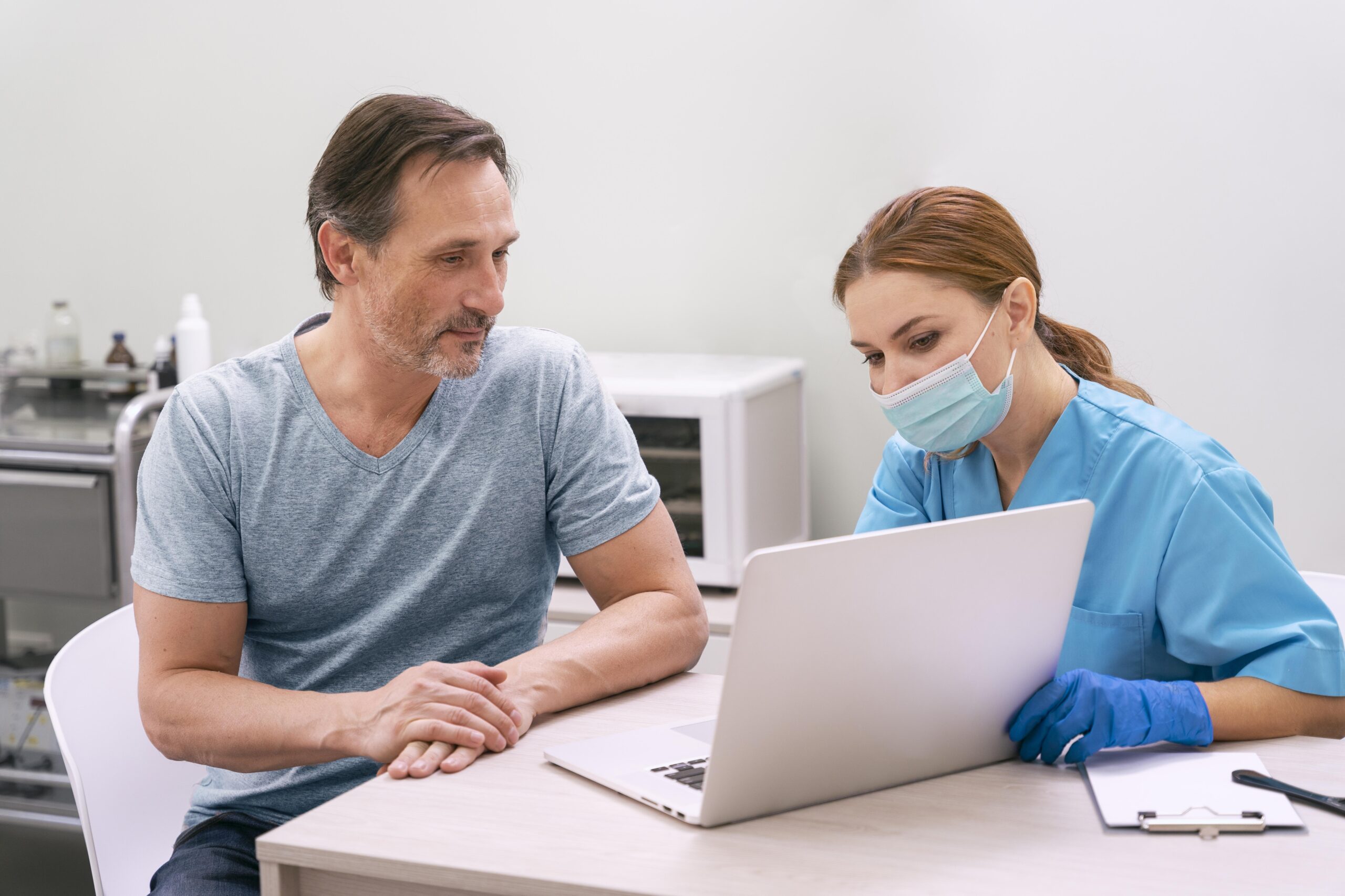 A healthcare provider in a blue uniform and face mask reviewing information on a laptop with a patient sitting beside her.