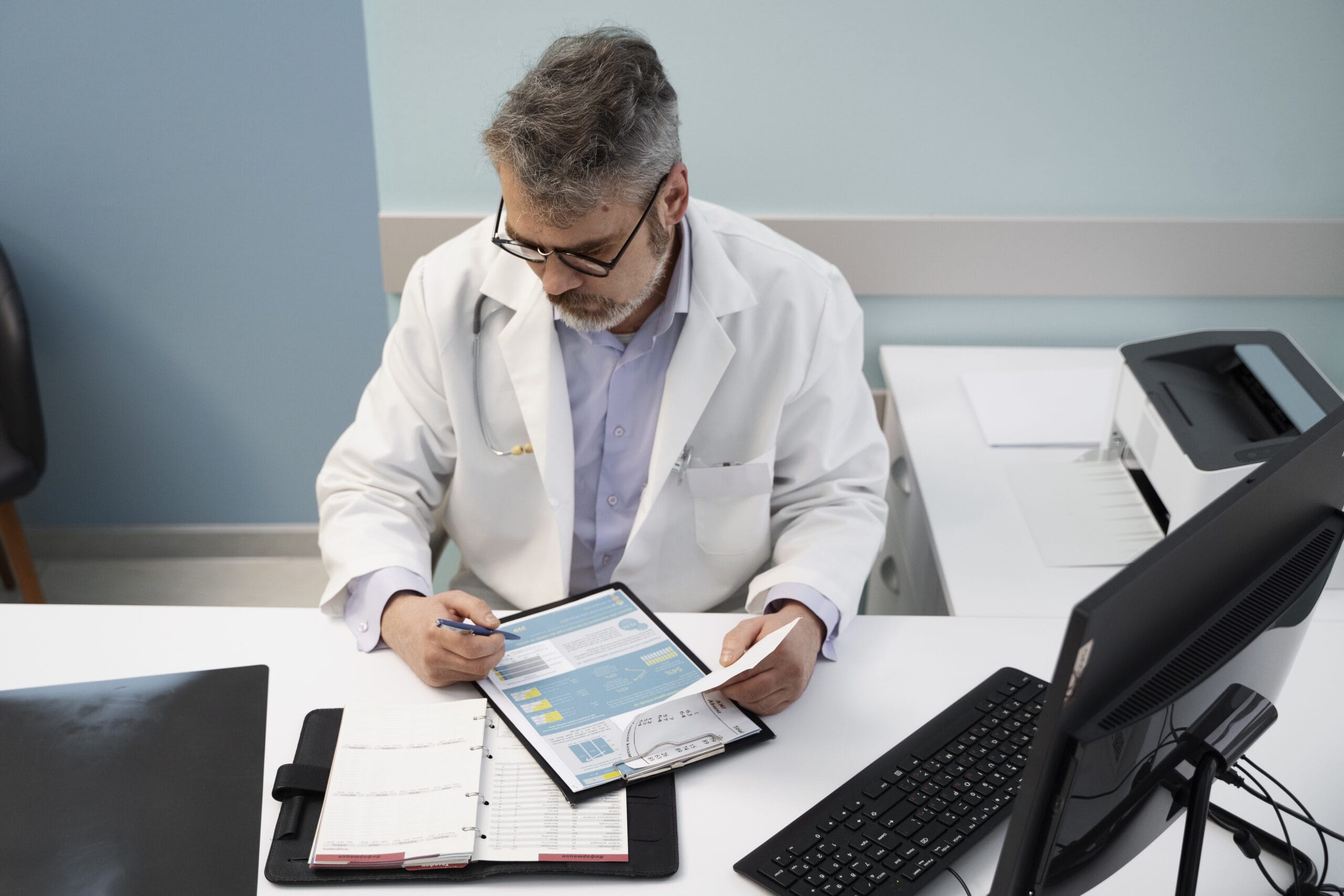 A healthcare provider reviewing documents at their desk, with a computer, a printed report, and a planner, symbolizing the prior authorization process in a medical setting.