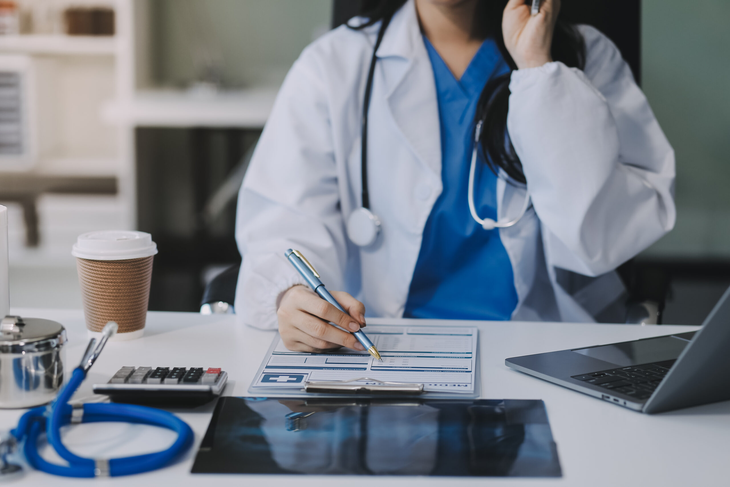 A healthcare provider in a rural clinic filling out medical forms with a stethoscope, laptop, and X-ray on the desk, symbolizing the importance of accurate medical coding for effective reimbursement and patient care.