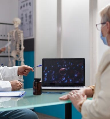 A doctor wearing a mask discussing a medical condition with a patient, showing diagnostic imaging on a laptop during the consultation.