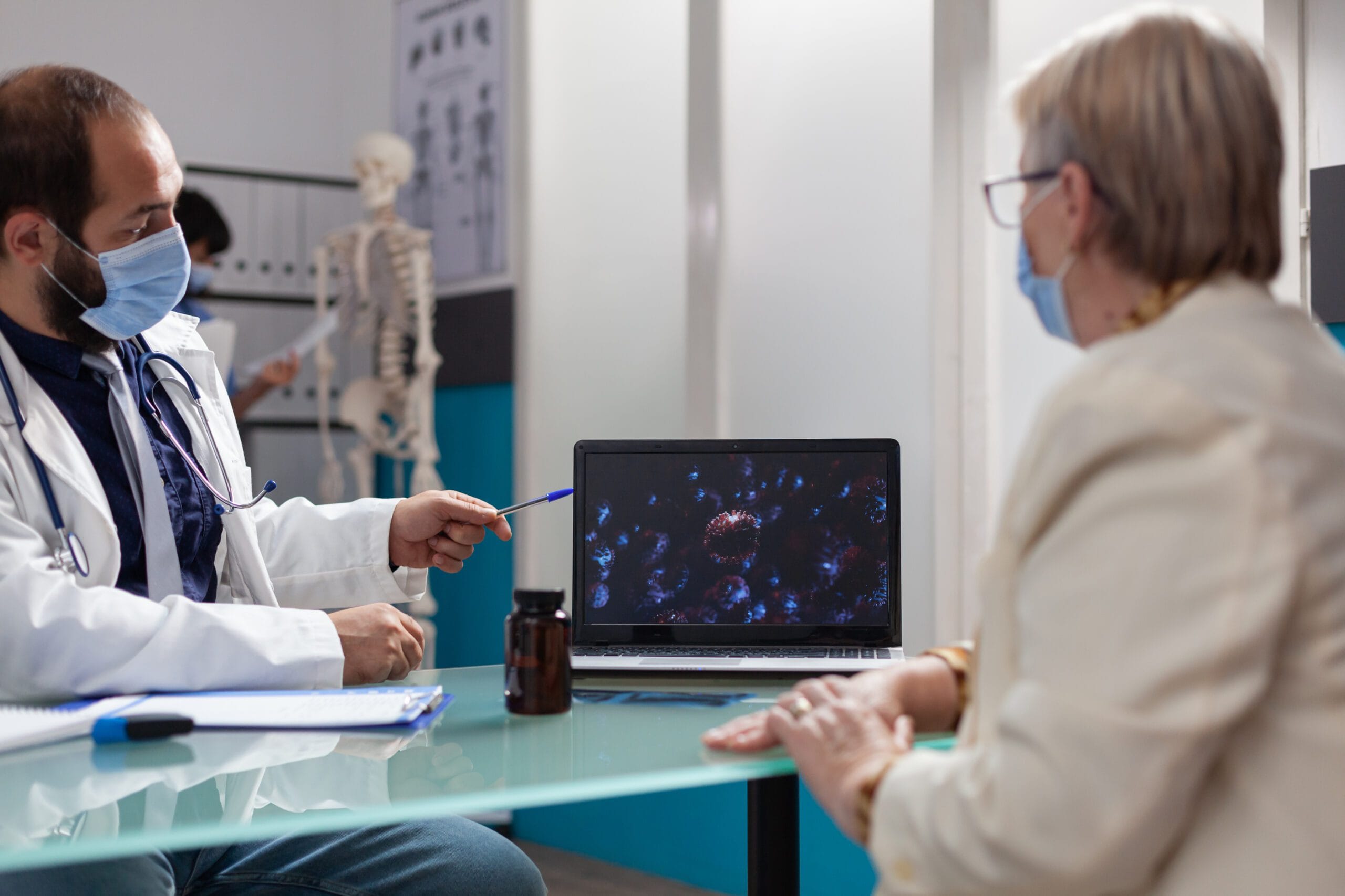 A doctor wearing a mask discussing a medical condition with a patient, showing diagnostic imaging on a laptop during the consultation.