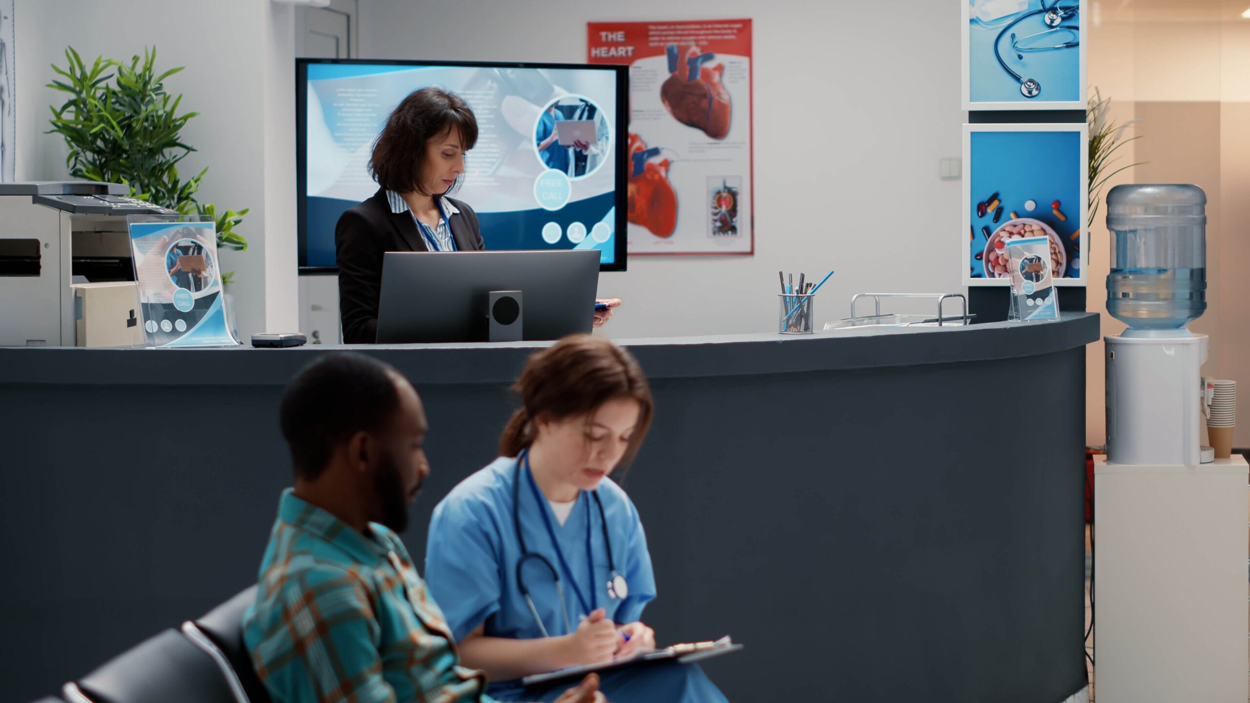 A hospital reception area with a receptionist working on a computer, a nurse reviewing patient files, and a patient waiting. The scene highlights administrative efficiency in healthcare settings.