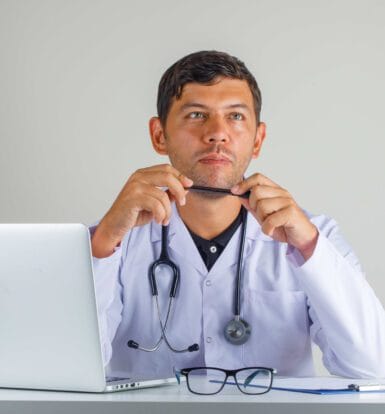 Doctor sitting thoughtfully at a desk with a laptop, stethoscope, and clipboard, representing telemedicine services and healthcare efficiency