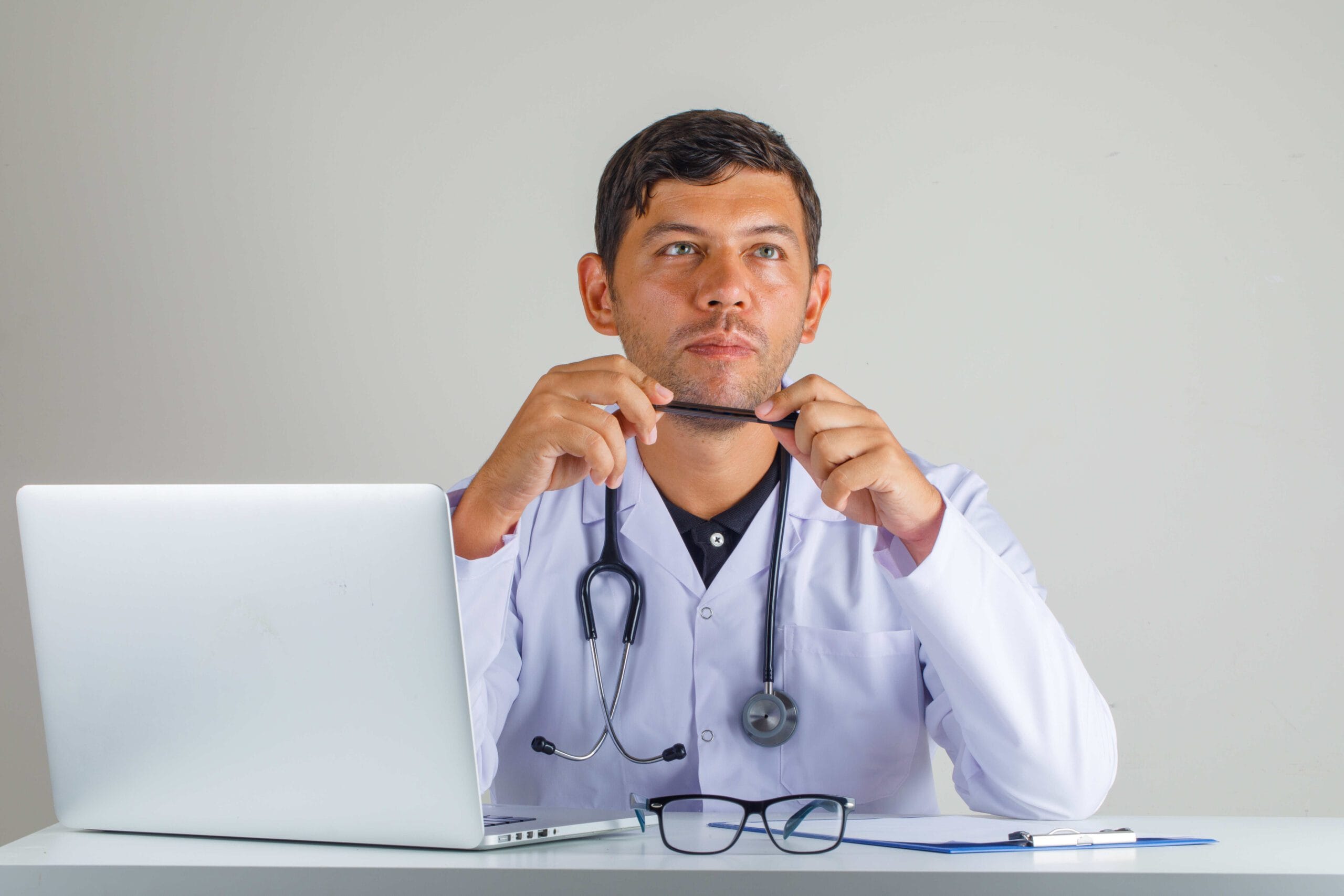 Doctor sitting thoughtfully at a desk with a laptop, stethoscope, and clipboard, representing telemedicine services and healthcare efficiency