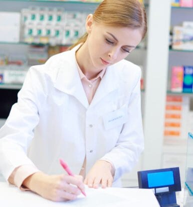 Pharmacist writing notes at a pharmacy counter with shelves of medications in the background.