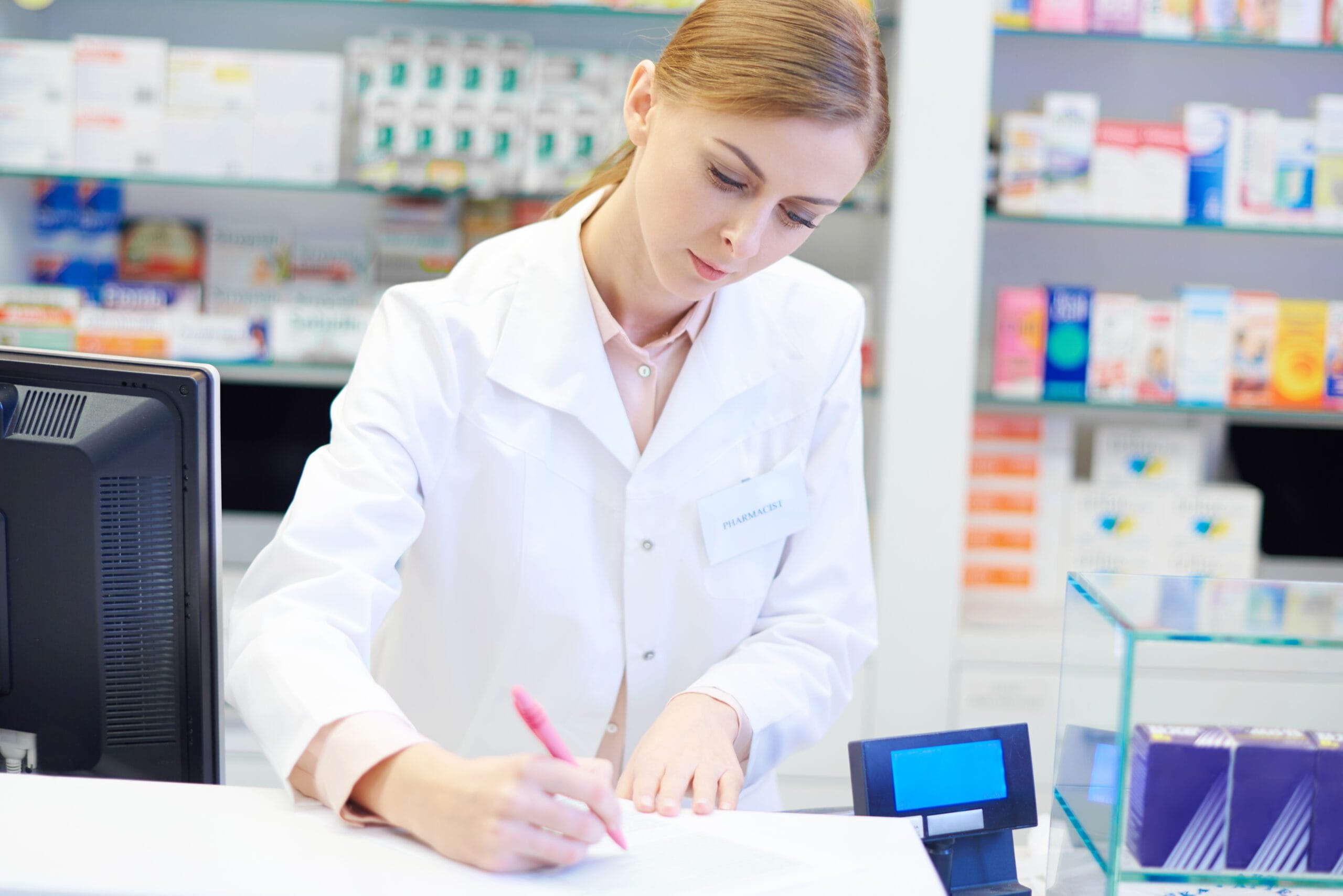 Pharmacist writing notes at a pharmacy counter with shelves of medications in the background.