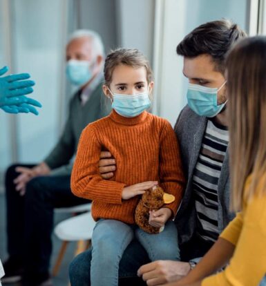 A healthcare provider wearing gloves discussing treatment options with a masked family, including a young girl holding a stuffed animal.