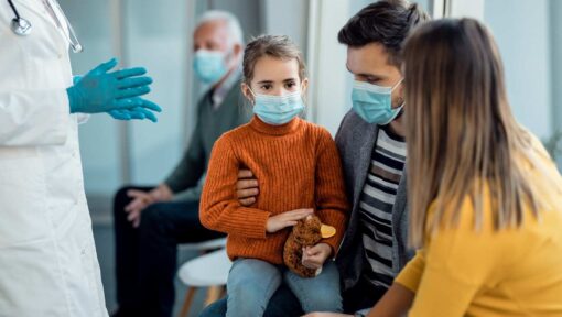 A healthcare provider wearing gloves discussing treatment options with a masked family, including a young girl holding a stuffed animal.