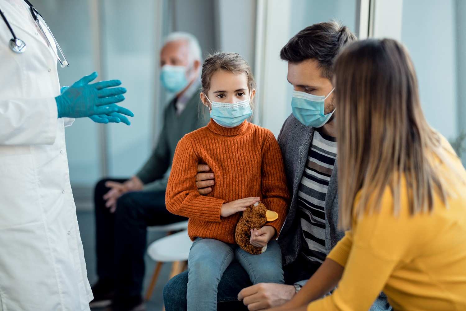 A healthcare provider wearing gloves discussing treatment options with a masked family, including a young girl holding a stuffed animal.