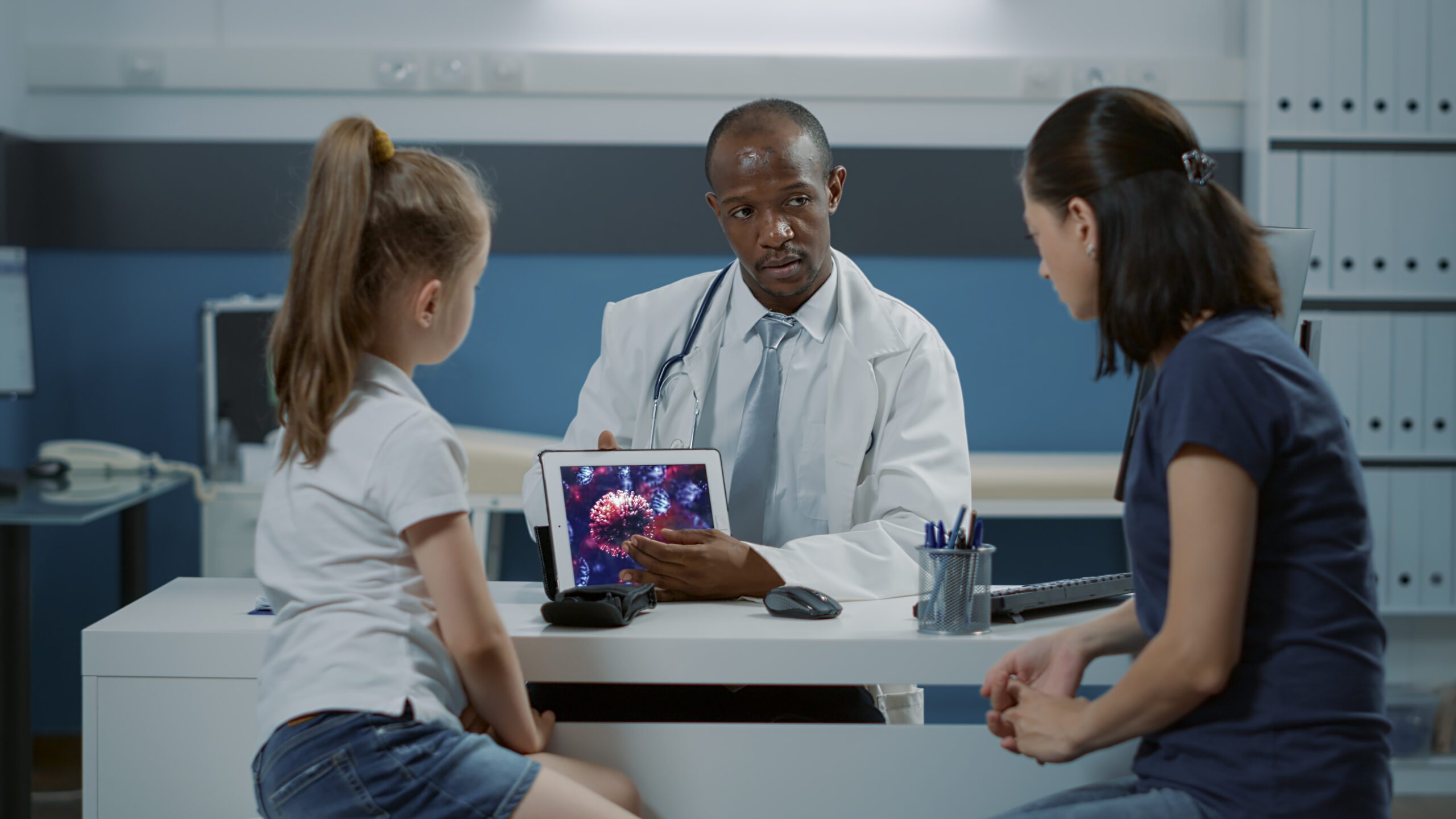 A doctor showing a digital image of a virus to a young patient and her mother, highlighting the use of medical technology in care.