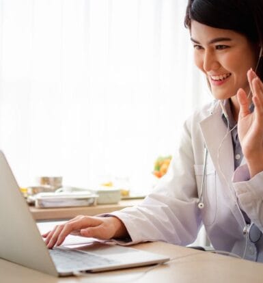 A smiling healthcare professional wearing earphones and interacting with a patient via a laptop, representing healthcare virtual assistant services.