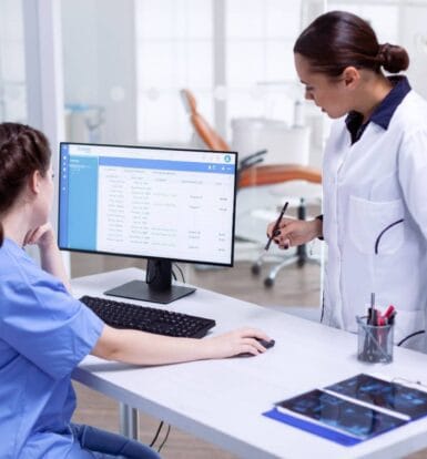 Two healthcare professionals reviewing patient records and codes on a computer in a clinical setting.