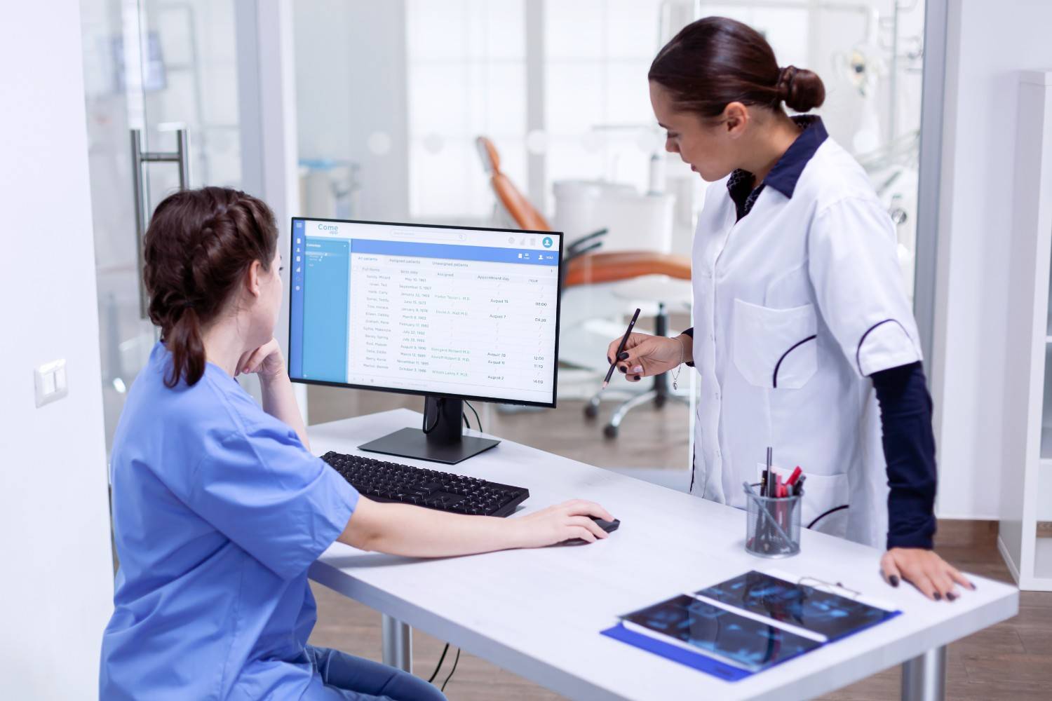 Two healthcare professionals reviewing patient records and codes on a computer in a clinical setting.