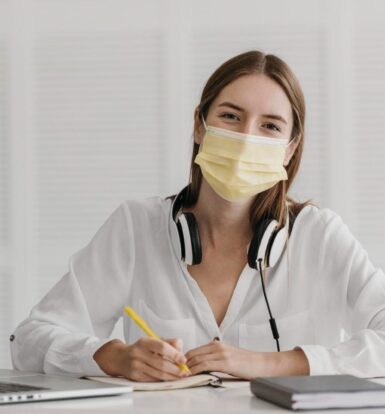 Young woman working remotely with headphones, mask, and laptop at a desk.