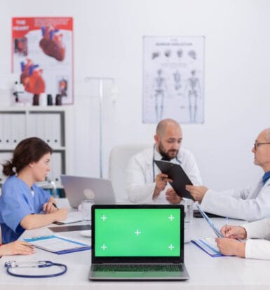 A team of doctors and nurses in a collaborative meeting, reviewing patient files and discussing medical strategies, with a laptop at the center symbolizing telemedicine support.