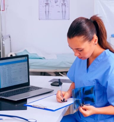 Healthcare professional in blue scrubs reviewing patient records and X-ray in a medical office with a laptop showing billing software on the screen.