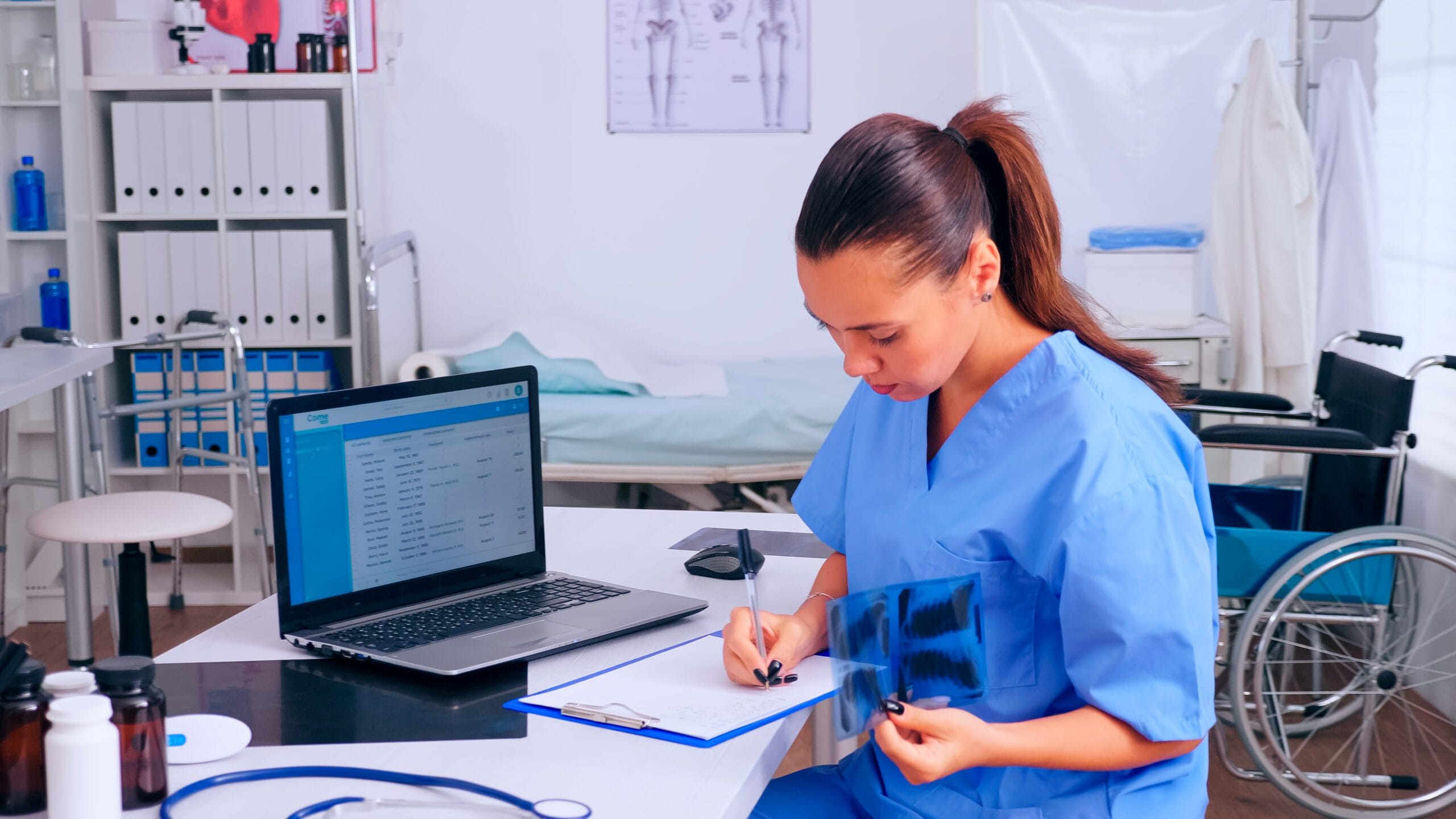 Healthcare professional in blue scrubs reviewing patient records and X-ray in a medical office with a laptop showing billing software on the screen.