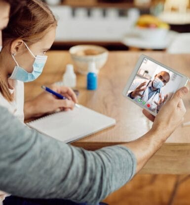 A father and child speaking with a masked doctor via telehealth on a tablet, showcasing secure and compliant telehealth services.