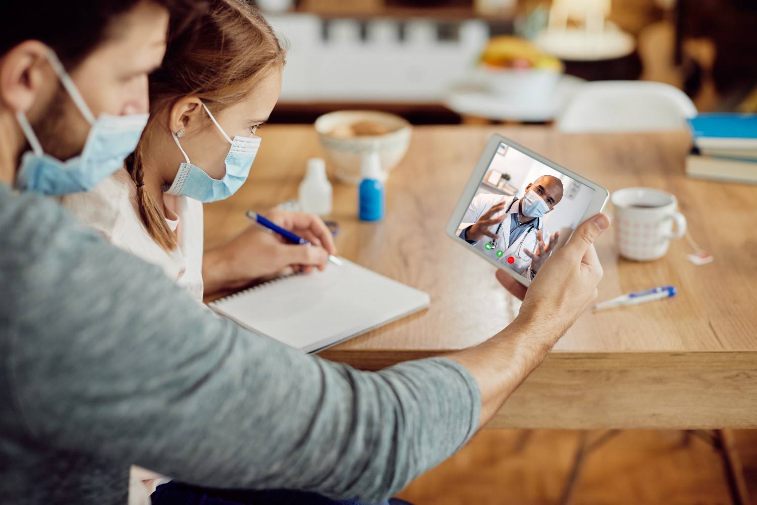 A father and child speaking with a masked doctor via telehealth on a tablet, showcasing secure and compliant telehealth services.