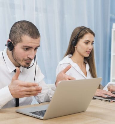 Two professional customer support representatives with headsets discussing telemedicine outsourcing solutions while working on laptops.