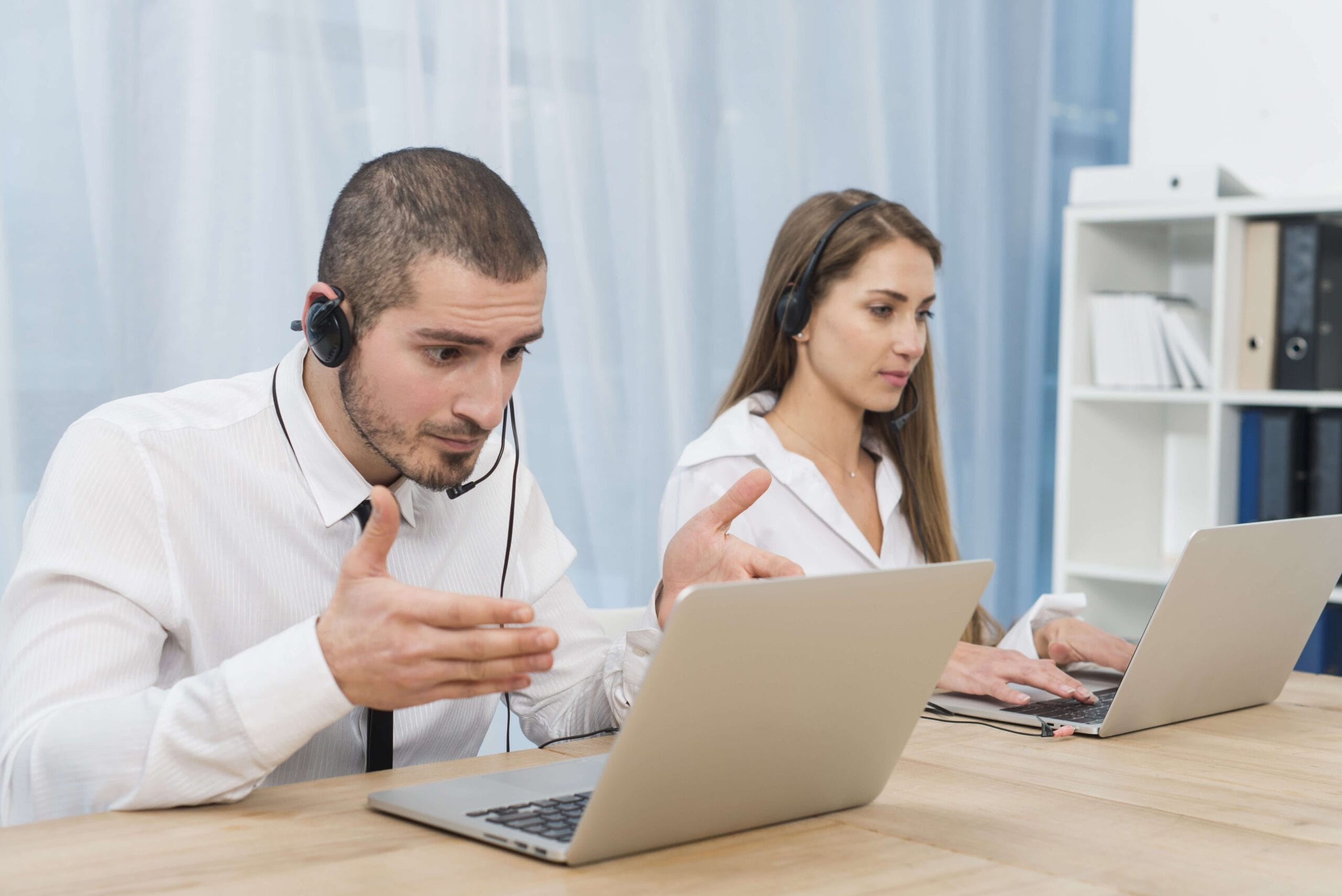 Two professional customer support representatives with headsets discussing telemedicine outsourcing solutions while working on laptops.