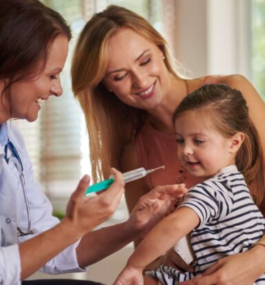 A smiling doctor holding a syringe while preparing to vaccinate a cheerful young girl, accompanied by her smiling mother.