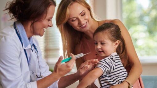 A smiling doctor holding a syringe while preparing to vaccinate a cheerful young girl, accompanied by her smiling mother.