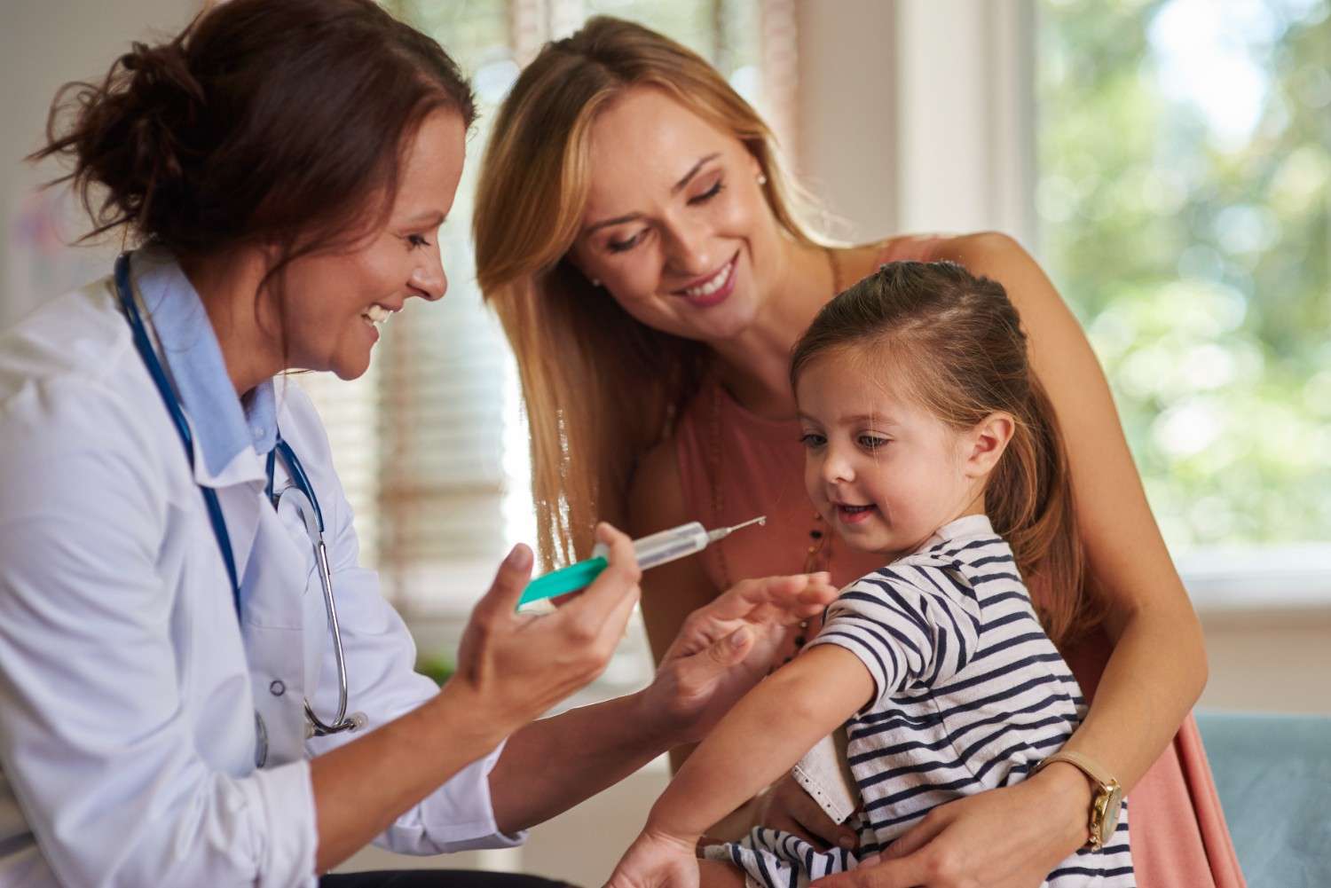 A smiling doctor holding a syringe while preparing to vaccinate a cheerful young girl, accompanied by her smiling mother.