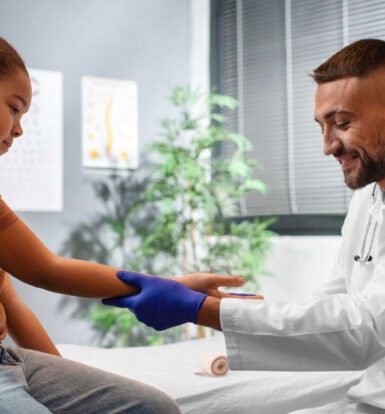 A doctor in a white coat and gloves is gently examining a young girl’s arm, smiling as they interact. The setting is a bright medical office with plants and a calm environment.